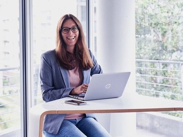 Lächelnde Frau mit langem braunen Haar in einem Büro sitzt vor ihrem Laptop.